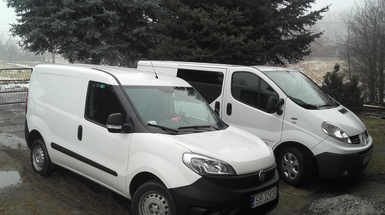 Dwa białe busy transportowe zaparkowane obok siebie na zewnątrz, z widocznymi numerami rejestracyjnymi i zabawką na desce rozdzielczej jednego z nich, na tle drzew i pochmurnego nieba.