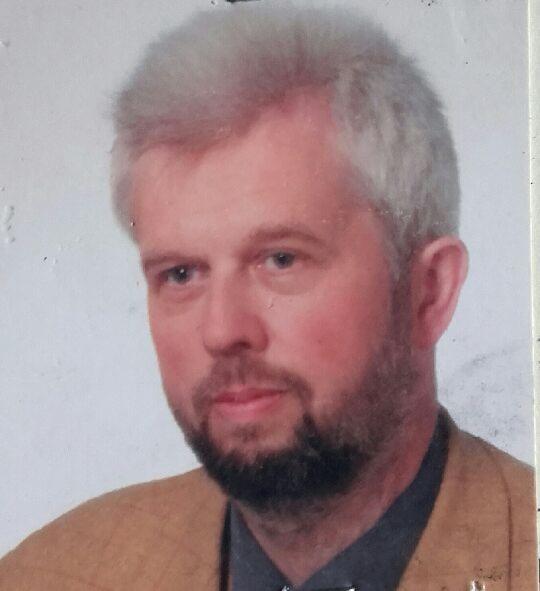 Portret mężczyzny z siwiejącymi włosami i brodą, ubrany w brązową marynarkę. Zdjęcie portretowe z delikatnym uśmiechem, wykonane w neutralnym oświetleniu.