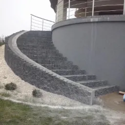 Zewnętrzne schody z szarego kamienia prowadzące na taras z metalową balustradą, otoczone białym żwirem i trawnikiem, widoczne ślady prac budowlanych.
