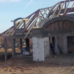 Surowy stan budowy domu z widoczną drewnianą konstrukcją dachu i elementami łukowymi nad otworami okiennymi, teren wokół domu nierówny i piaszczysty.