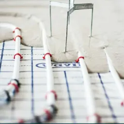 Świeżo wylana, jasnoszara masa na ogrzewanie podłogowe z białymi rurkami i czerwonymi uchwytami, widoczne metalowe podpory do poziomowania w tle.