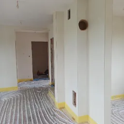 Wnętrze w trakcie prac wykończeniowych: świeżo położone tynki maszynowe na ścianach oraz instalacja ogrzewania podłogowego z rurkami na siatce, zabezpieczone folią przy ścianach.