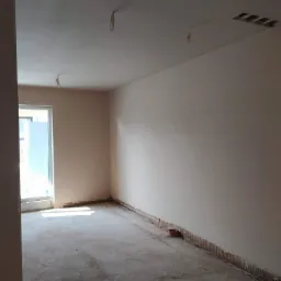 Surowe wnętrze pokoju z otynkowanymi ścianami, betonową posadzką przed wylewką i wystającymi przewodami elektrycznymi z sufitu.