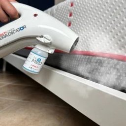 Odpluskwianie-24 - Biały parowy aplikator z pojemnikiem na płyn, rozpyla preparat na materac łóżka z czerwoną lamówką. Widoczna para wydobywająca się z aplikatora.