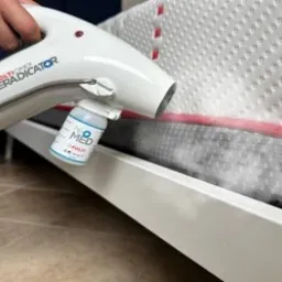 Biały parowy aplikator z pojemnikiem na płyn, rozpyla preparat na materac łóżka z czerwoną lamówką. Widoczna para wydobywająca się z aplikatora.