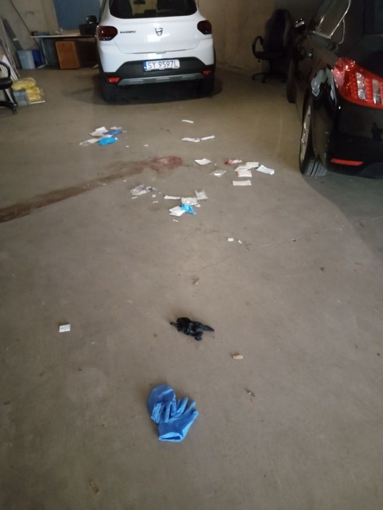Podłoga garażu z plamą przypominającą krew, porozrzucanymi papierami, niebieską rękawiczką lateksową i czarnym przedmiotem, pomiędzy zaparkowanymi samochodami osobowymi.