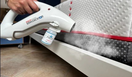 Biały parowy aplikator z pojemnikiem na płyn, rozpyla preparat na materac łóżka z czerwoną lamówką. Widoczna para wydobywająca się z aplikatora.