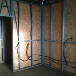 Wnętrze w trakcie budowy z widoczną metalową konstrukcją ścian działowych, płytami OSB i poprowadzonymi przewodami elektrycznymi wewnątrz ścian.