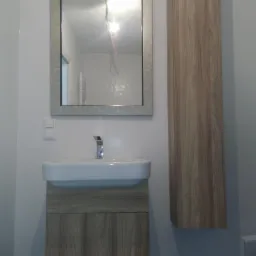 Nowoczesna łazienka z szafką pod umywalką z drewnopodobnym frontem, lustrem w szarej ramie z oświetleniem LED i wysoką szafką wiszącą z forniru.