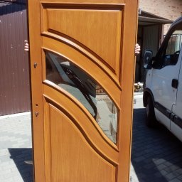 Zakład Stolarski Wojciech Krawiec - Lakierowane drzwi z szybą o falistym wzorze, trzymane przez dwie osoby na zewnątrz, w tle budynek i biały samochód dostawczy. Widoczne detale wykończenia i okucia.