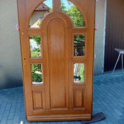 Zakład Stolarski Wojciech Krawiec - Brązowe, drewniane drzwi z szybami, częściowo zasłonięte przez dwie osoby. Widoczne odbicie otoczenia w szybach. Drzwi stoją na kostce brukowej.