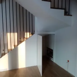 Nowoczesne schody z drewnianymi stopniami i czarną metalową balustradą, z ukrytym pomieszczeniem pod biegiem schodów i oświetleniem słonecznym padającym na konstrukcję.