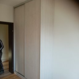 Zakład Stolarski Wojciech Krawiec - Jasna szafa przesuwna z drewnopodobnymi frontami, zamontowana w rogu pokoju z panelami podłogowymi, widoczny fragment osoby w drzwiach.