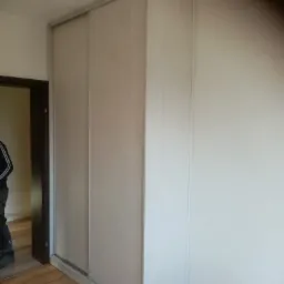 Jasna szafa przesuwna z drewnopodobnymi frontami, zamontowana w rogu pokoju z panelami podłogowymi, widoczny fragment osoby w drzwiach.