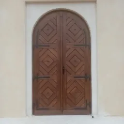 Dwuskrzydłowe, drewniane wejściowe z łukowym zwieńczeniem i dekoracyjnym wzorem rombów, osadzone w jasnej ścianie.