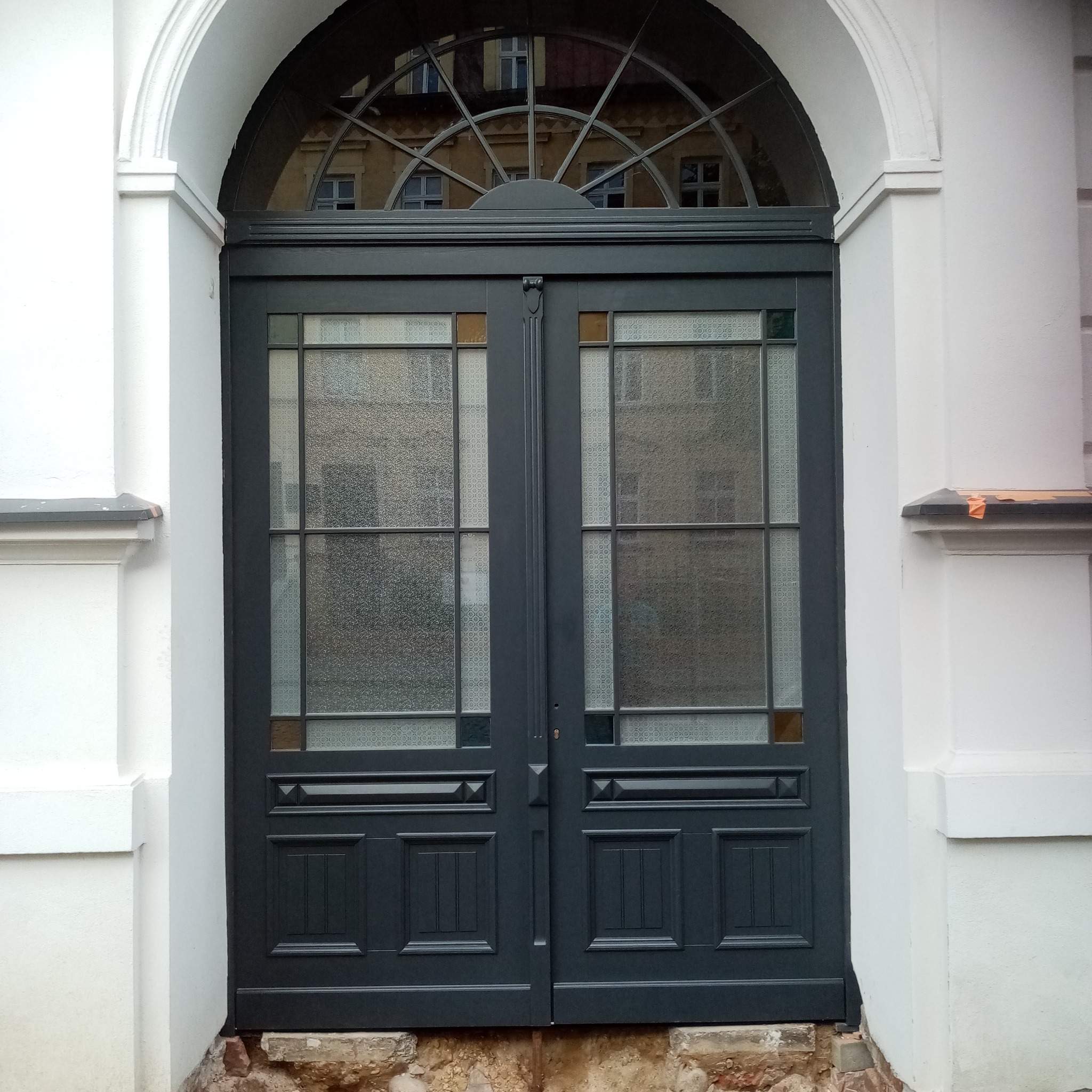 Eleganckie, grafitowe drzwi dwuskrzydłowe z dekoracyjnymi szybami i łukowym nadświetlem, wkomponowane w białą fasadę budynku. Widoczne detale architektoniczne.