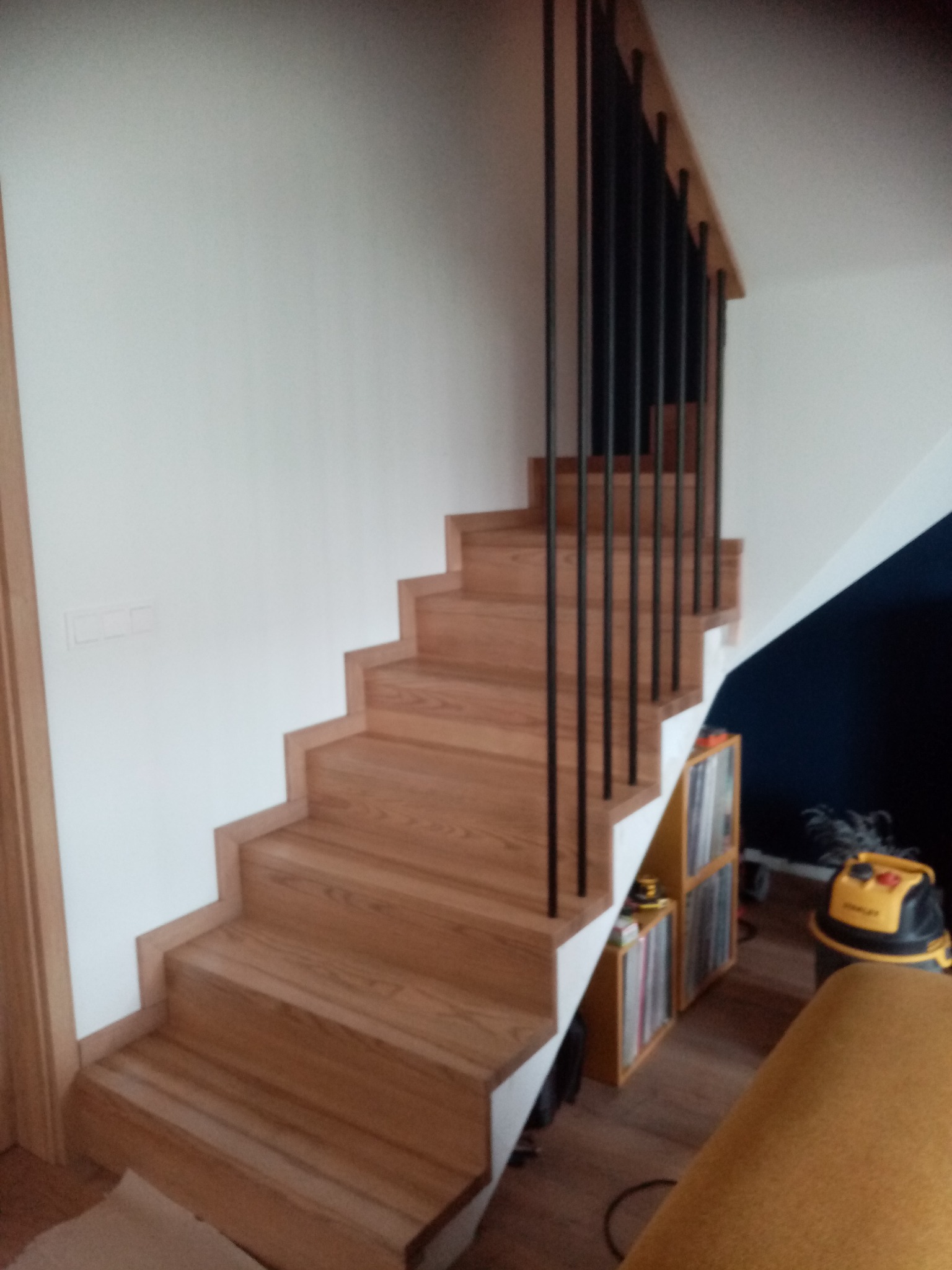 Drewniane schody z czarną, metalową balustradą w nowoczesnym wnętrzu. Widoczne stopnie z jasnego drewna i białe ściany. W tle regał i odkurzacz.