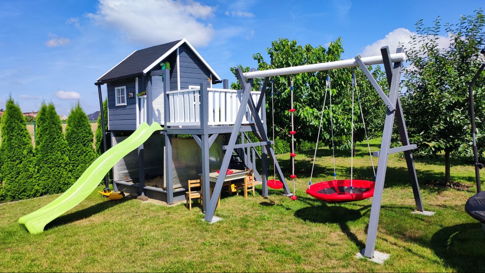 Drewniany plac zabaw z domkiem, zjeżdżalnią i huśtawką na tle zielonego trawnika i błękitnego nieba. Widoczne detale: numer domu '79', stół z krzesłami pod domkiem.