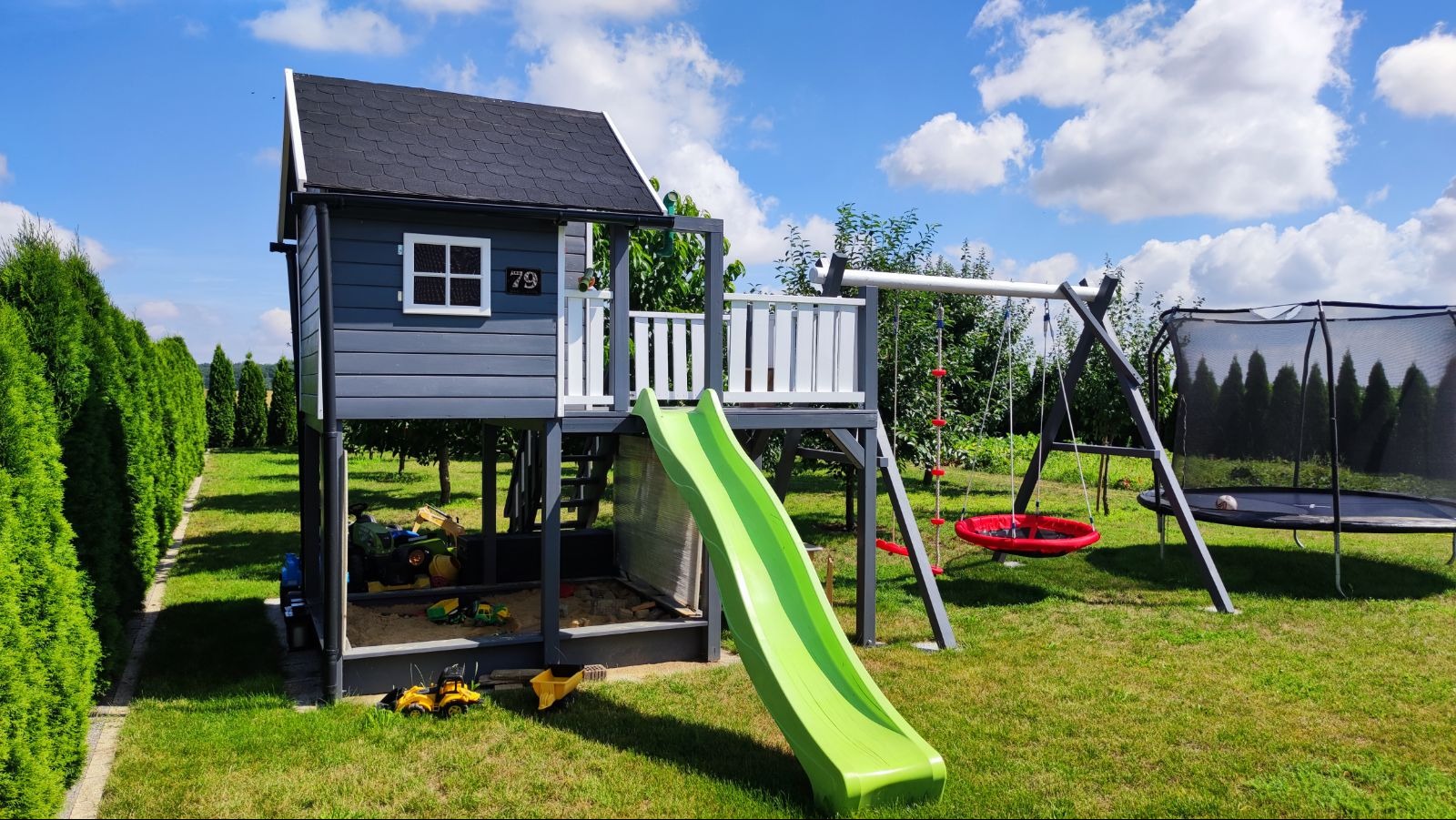 Drewniany plac zabaw dla dzieci z domkiem, zjeżdżalnią i huśtawką na tle zielonego trawnika i błękitnego nieba z chmurami. Widoczne zabawki w piaskownicy.
