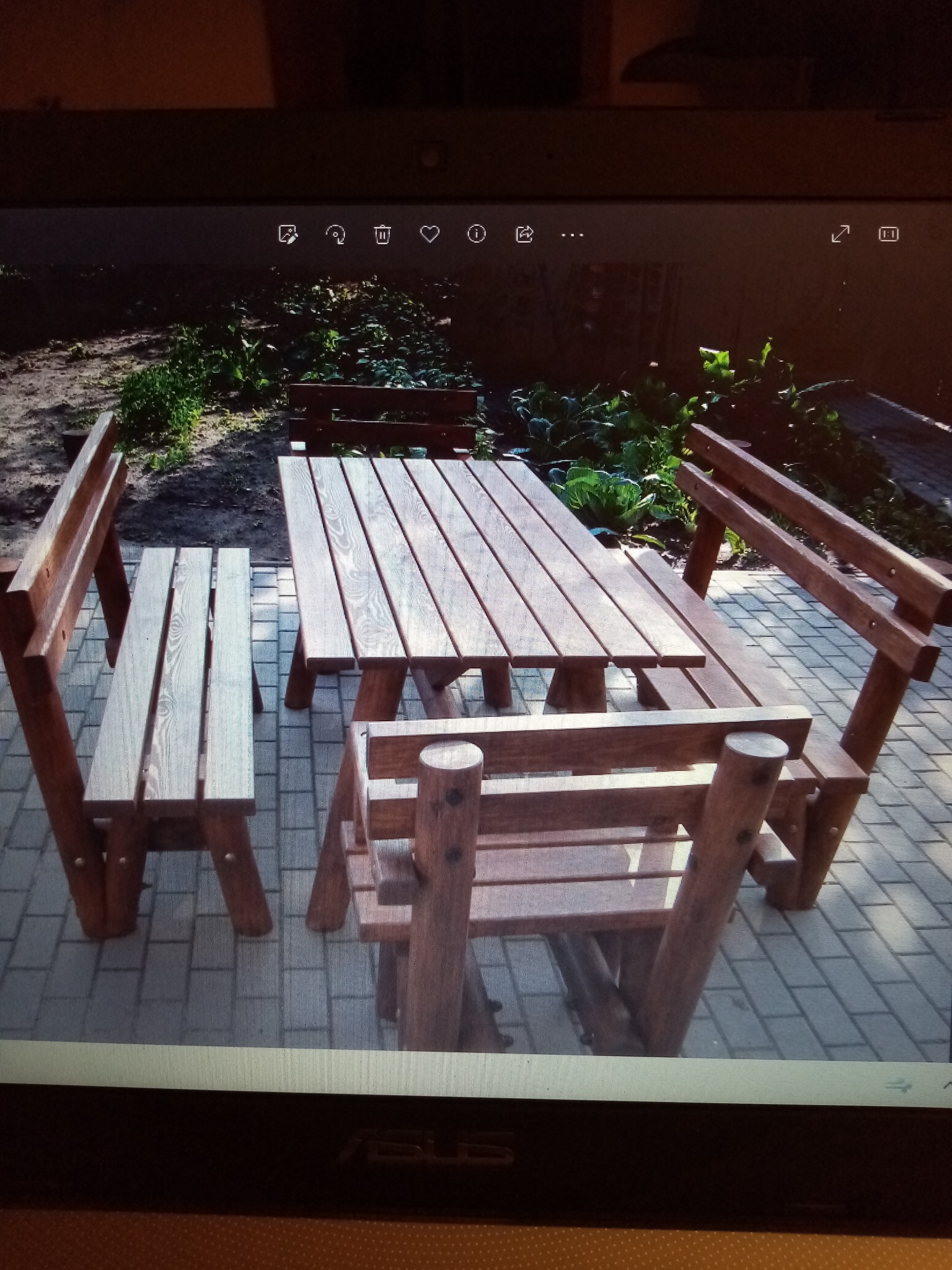 Drewniany stół piknikowy z czterema ławkami na brukowanym patio, otoczony zielenią. Widoczne słoje drewna i solidna konstrukcja zestawu.