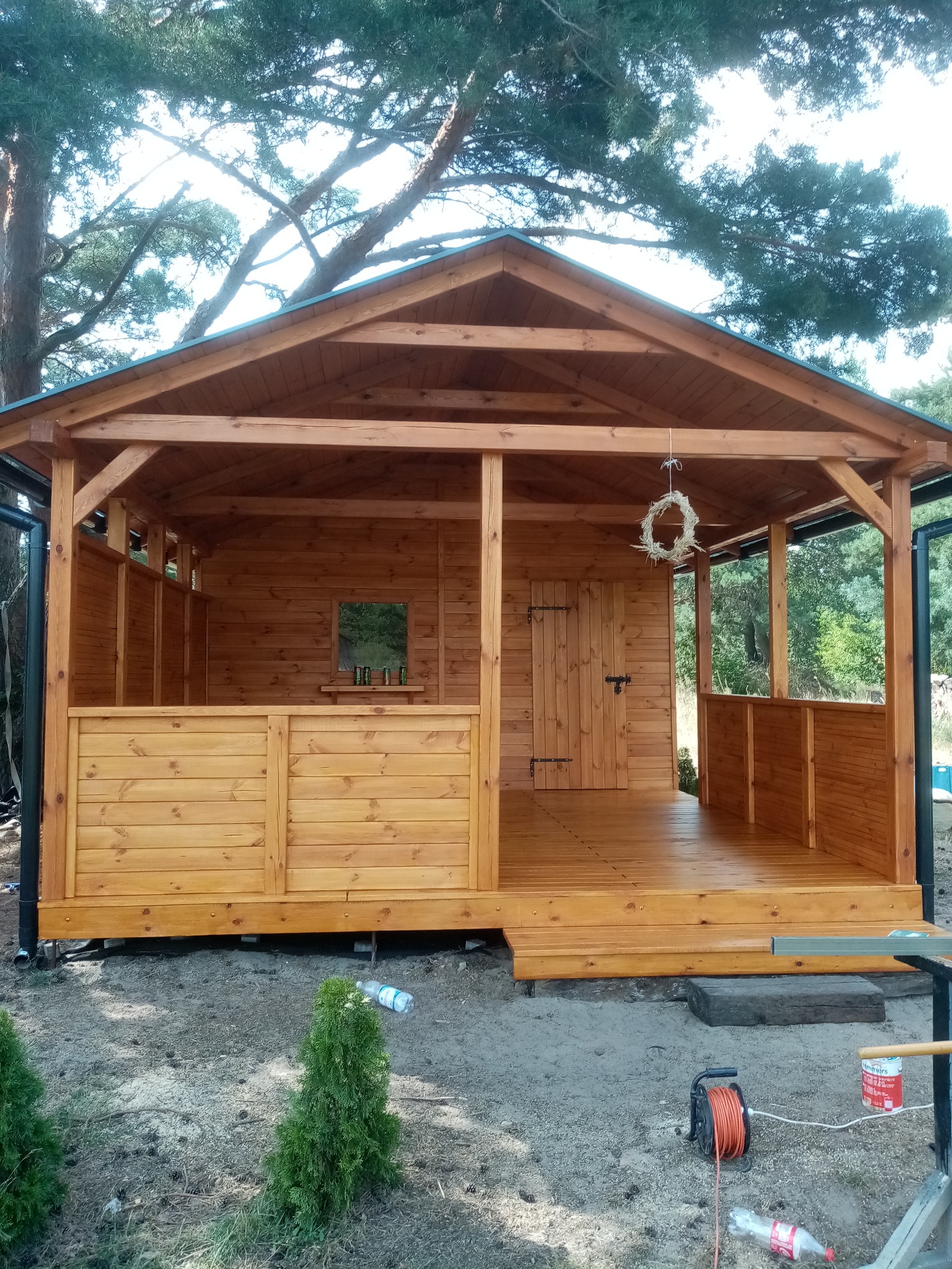 Drewniana altana ogrodowa z zadaszeniem, widoczne detale konstrukcyjne, drzwi z zawiasami i zasuwa, wieniec dekoracyjny, butelki na półce. Otoczenie naturalne, drzewa w tle.