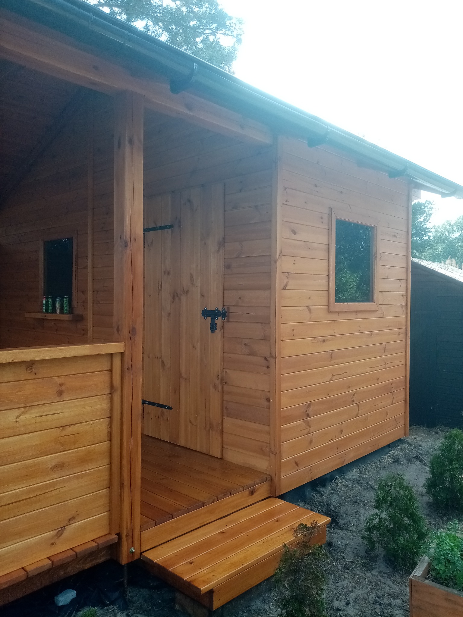 Drewniana altana ogrodowa z zadaszeniem, widoczne drzwi z czarnym zamkiem, okno i schodki wejściowe. Konstrukcja z jasnego drewna, otoczona zielenią.