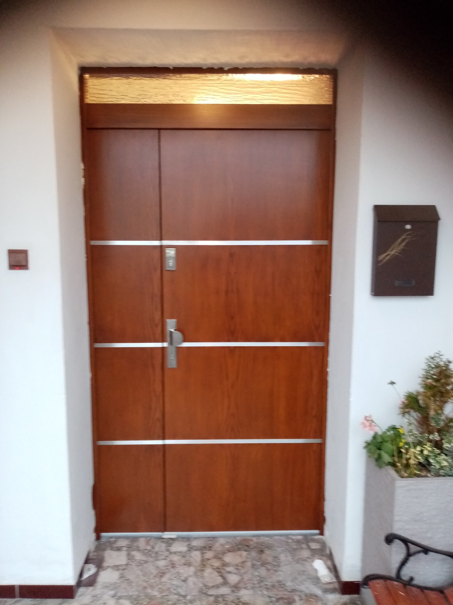 Brązowe drzwi wejściowe z przeszkleniem u góry i trzema poziomymi, srebrnymi paskami, zamontowane w białej ścianie z brązową skrzynką na listy.