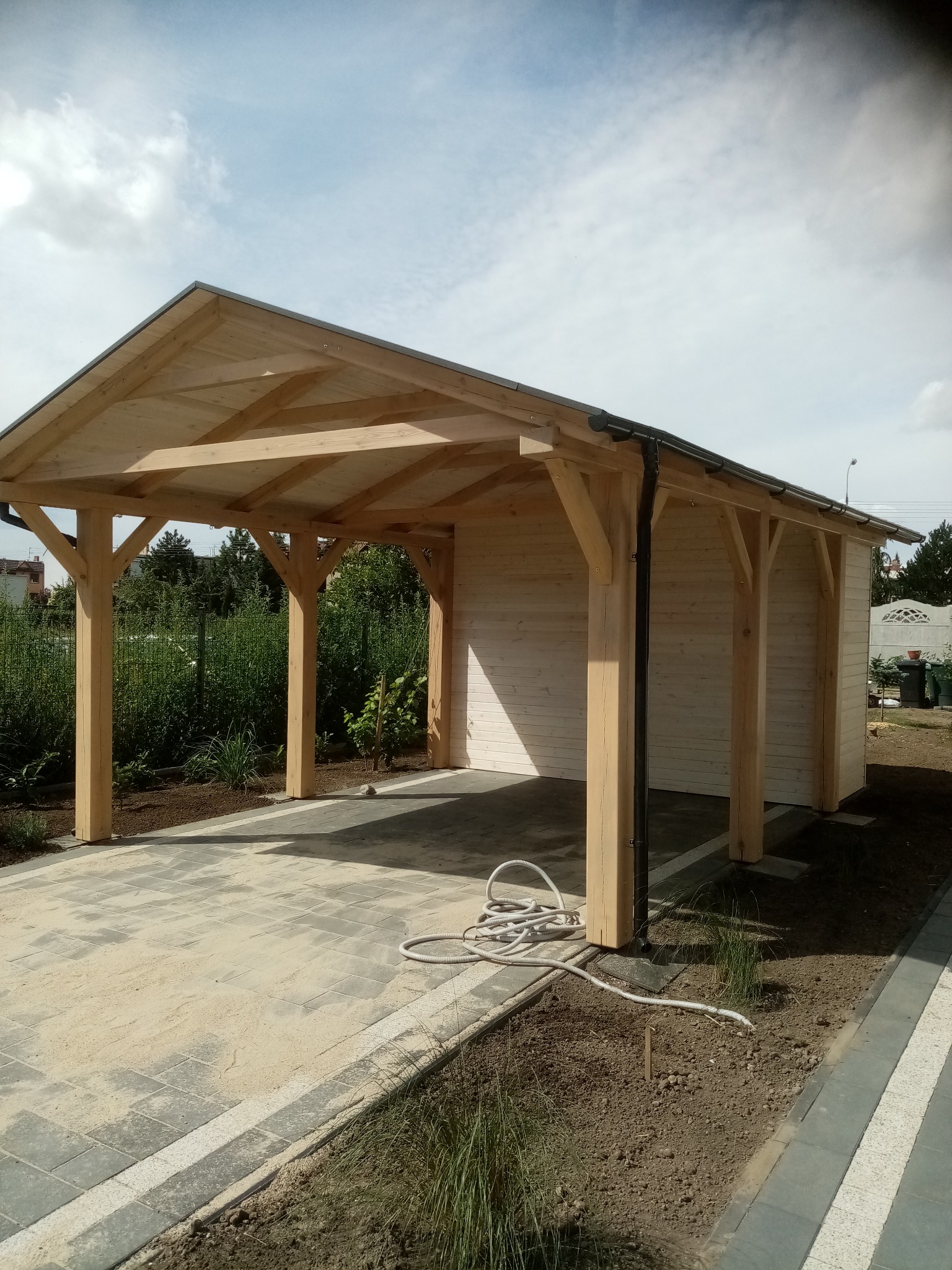 Drewniana wiata garażowa z jasnego drewna, z widoczną konstrukcją dachu i słupami. Na ziemi kostka brukowa i zwinięty wąż ogrodowy. W tle zieleń i zabudowania.