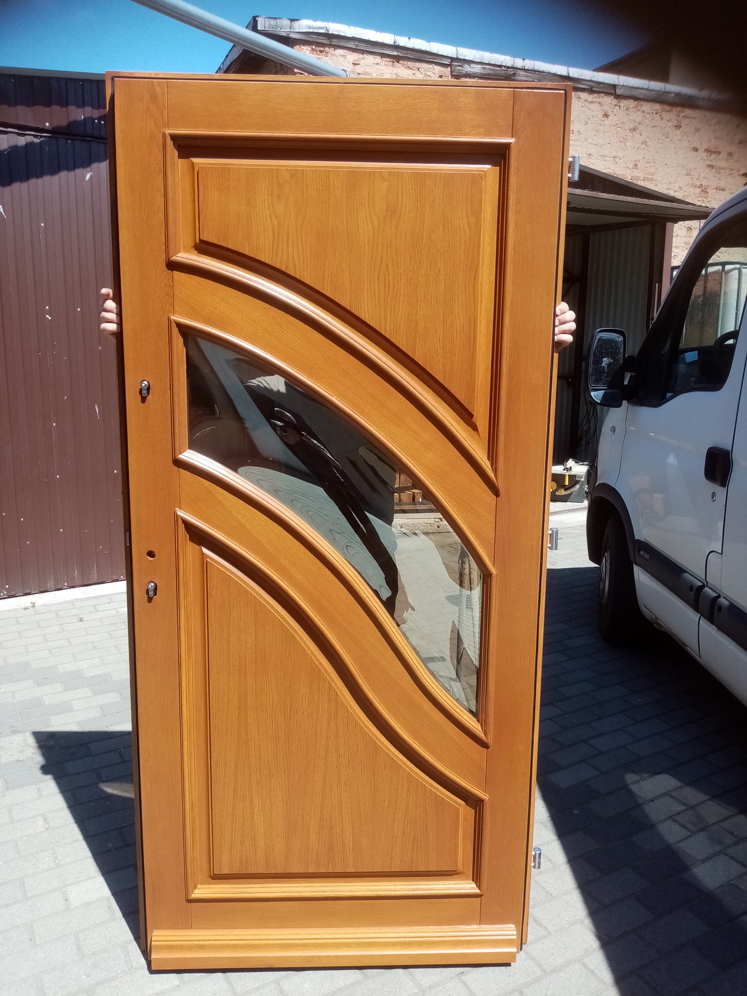 Lakierowane drzwi z szybą o falistym wzorze, trzymane przez dwie osoby na zewnątrz, w tle budynek i biały samochód dostawczy. Widoczne detale wykończenia i okucia.
