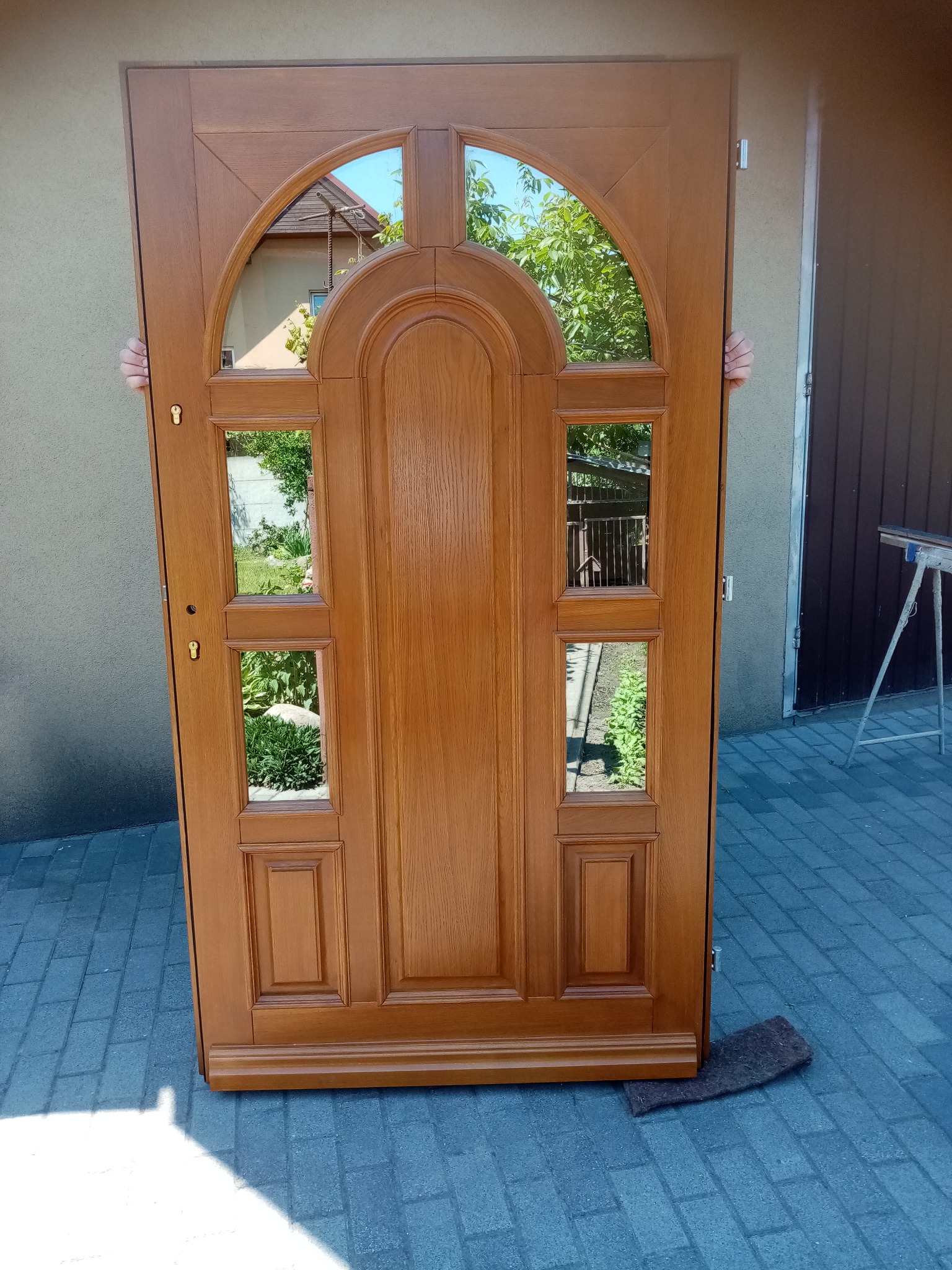 Brązowe, drewniane drzwi z szybami, częściowo zasłonięte przez dwie osoby. Widoczne odbicie otoczenia w szybach. Drzwi stoją na kostce brukowej.