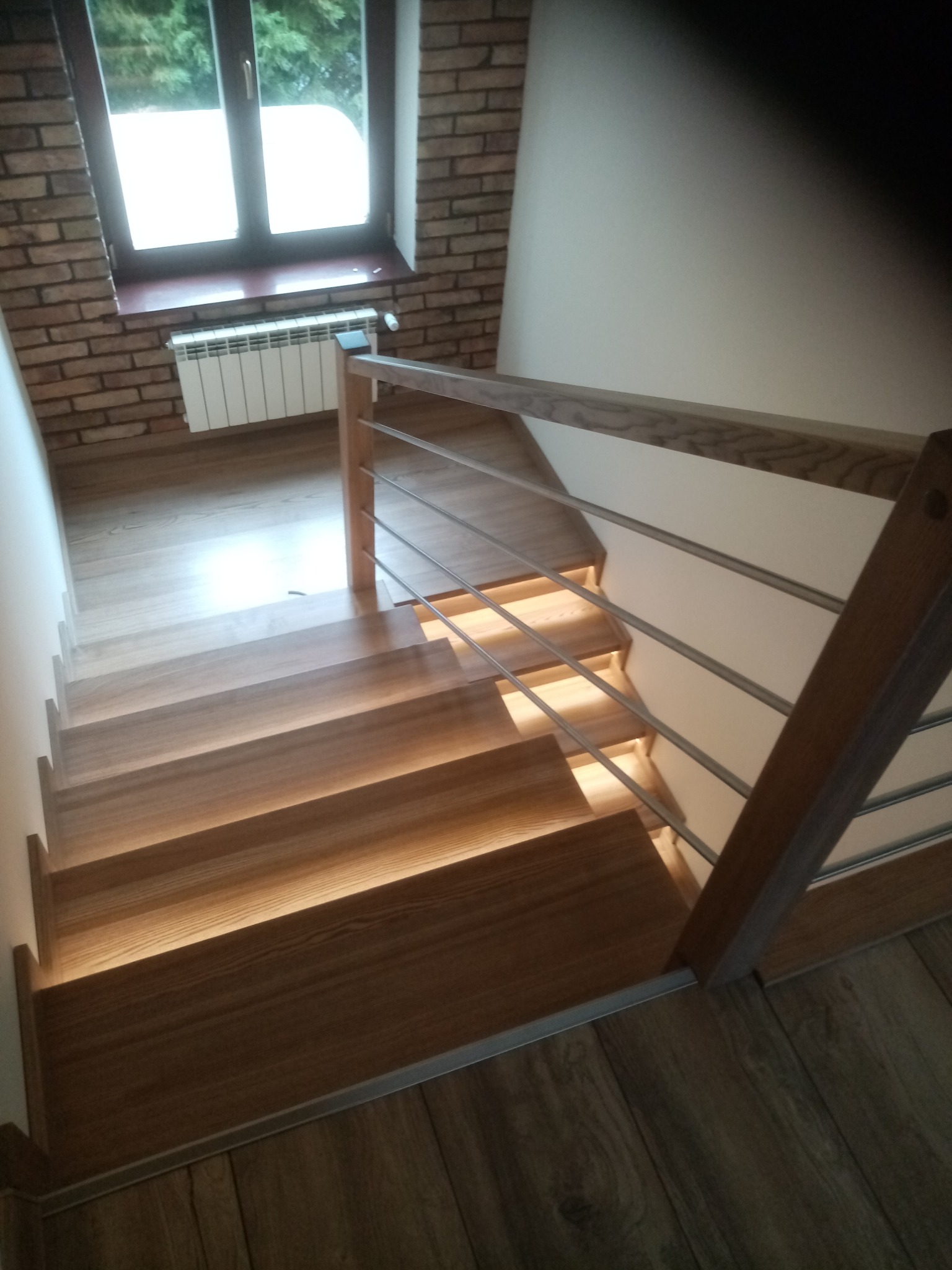 Nowoczesne schody drewniane z oświetleniem LED, metalowe barierki, widok z góry. Naturalne drewno, ceglana ściana z oknem w tle. Detal wykończenia.