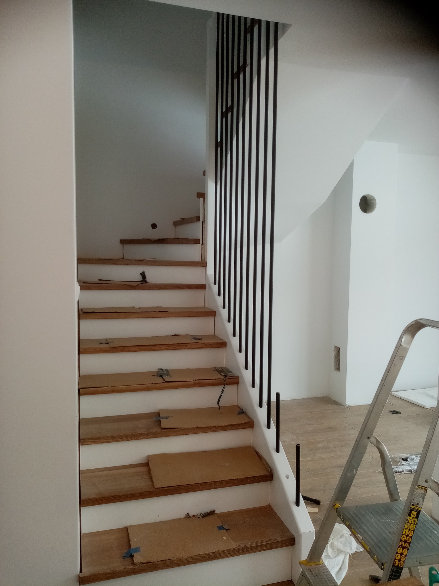 Wnętrze domu w trakcie wykańczania: drewniane schody z białymi podstopnicami i czarną balustradą, zabezpieczone kartonem, drabina w tle. Etap prac wykończeniowych.
