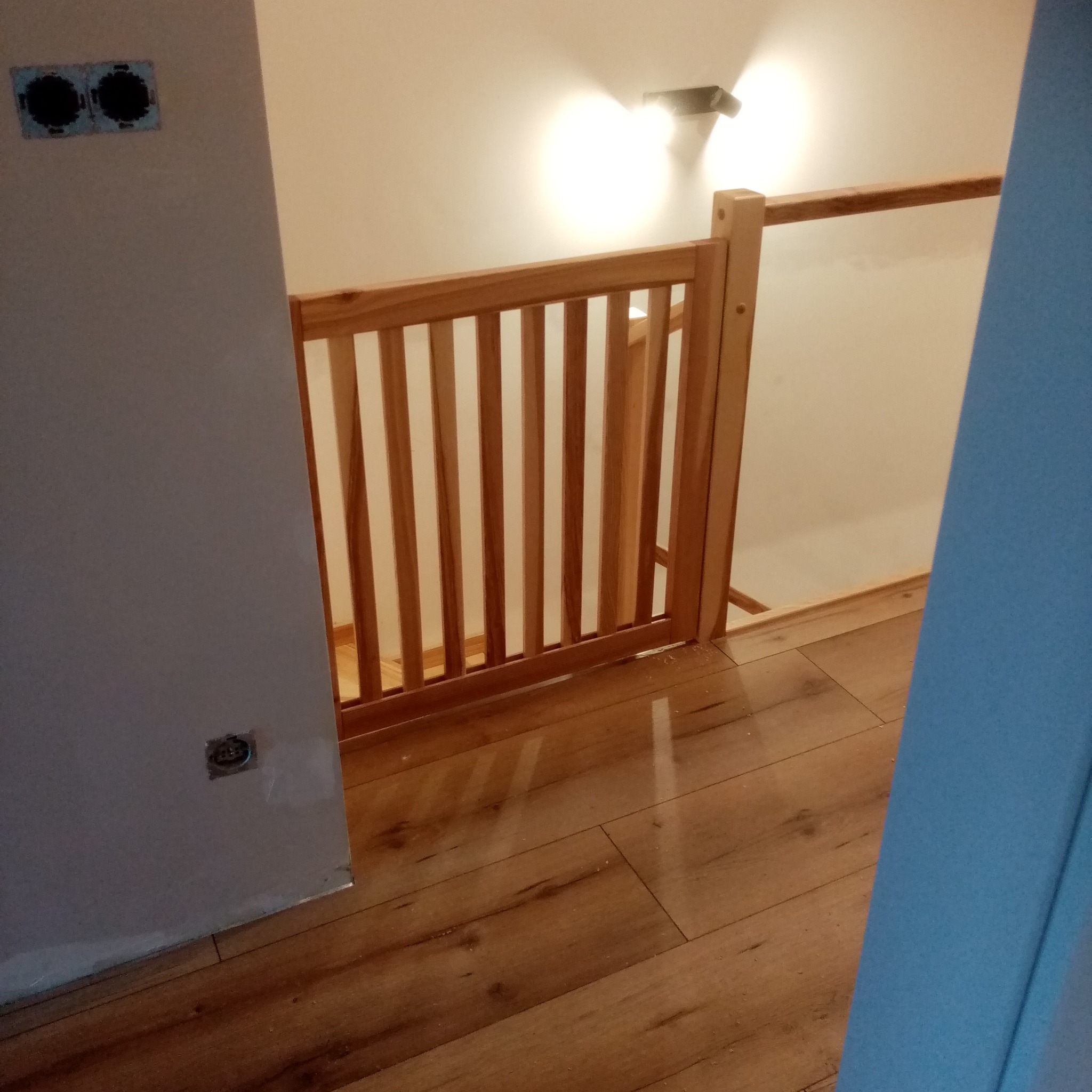 Jasne, drewniane zabezpieczenie schodów z pionowymi szczebelkami, zamontowane na drewnianej podłodze. Widoczne gniazdka elektryczne na ścianie.