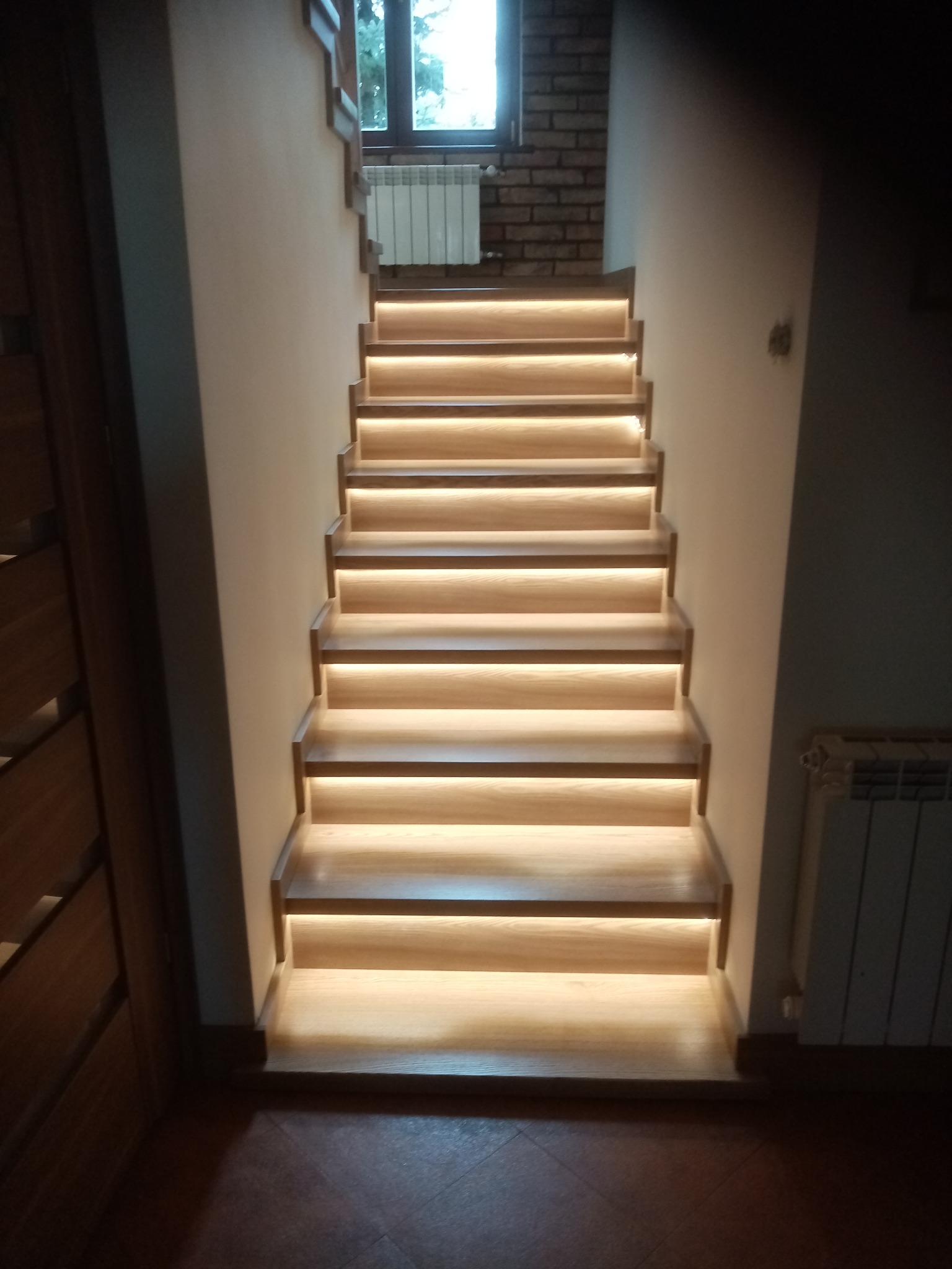Nowoczesne schody drewniane z podświetleniem LED, widok z dołu. Stopnie z jasnego drewna, ciepłe światło. Ujęcie centralne, widoczny fragment okna i grzejnika na podeście.