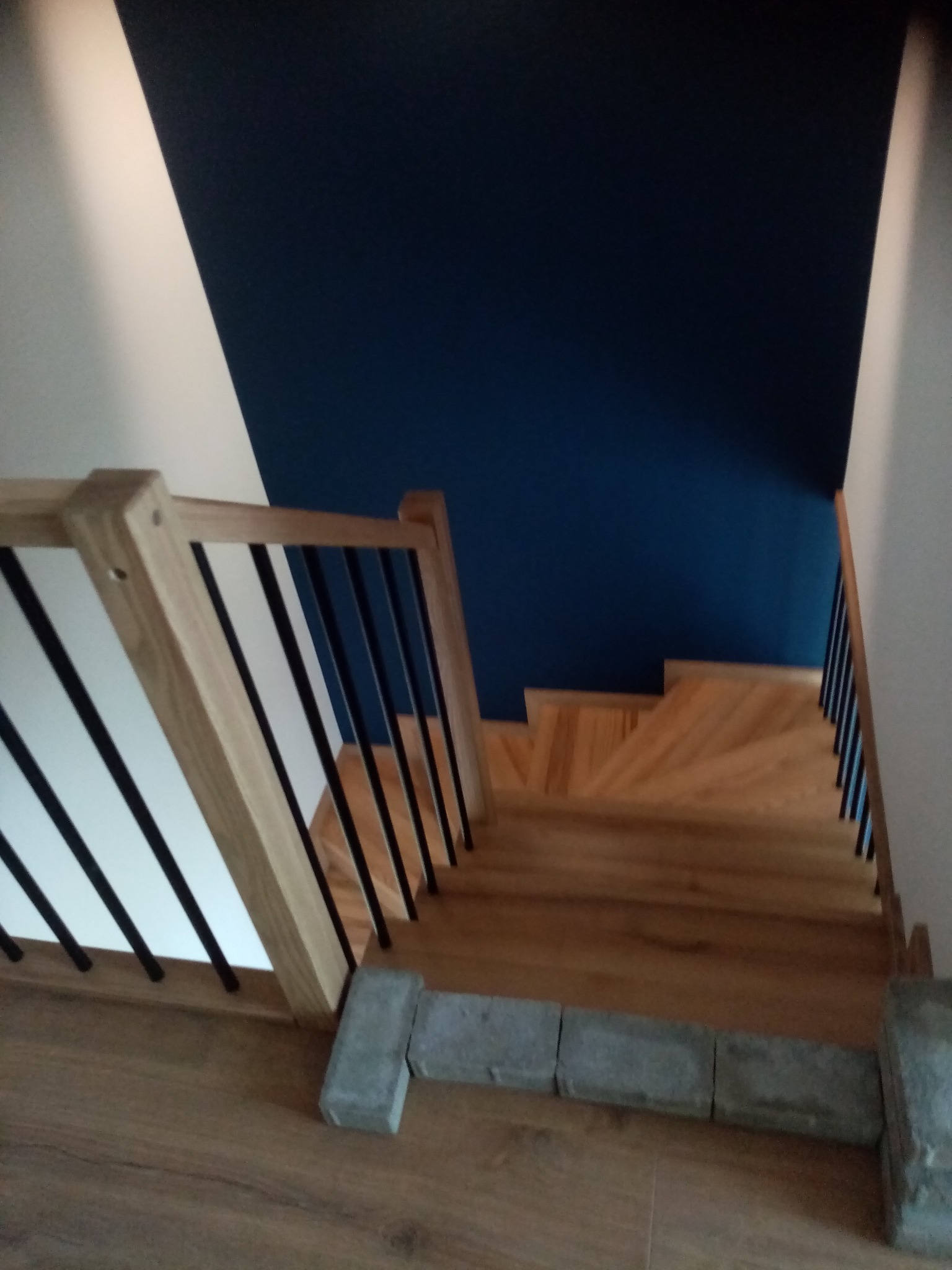 Jasne, drewniane schody z czarnymi balustradami na tle ściany w kolorze granatowym i białym. Widok z góry, ukazujący detale wykończenia i nowoczesny design.