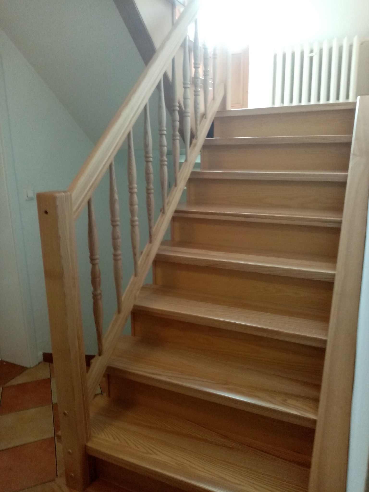 Jasne, drewniane schody z ozdobną balustradą wewnątrz domu. Widoczne stopnie i fragment podłogi z terakoty. W tle grzejnik.