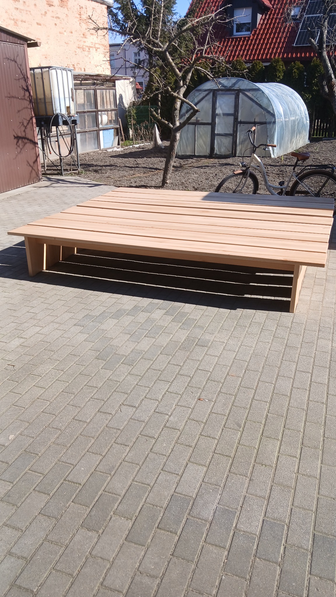 Drewniany, niski stół ogrodowy z desek na kostce brukowej, w tle szklarnia i rower. Jasne drewno, słoneczny dzień, widok z boku.