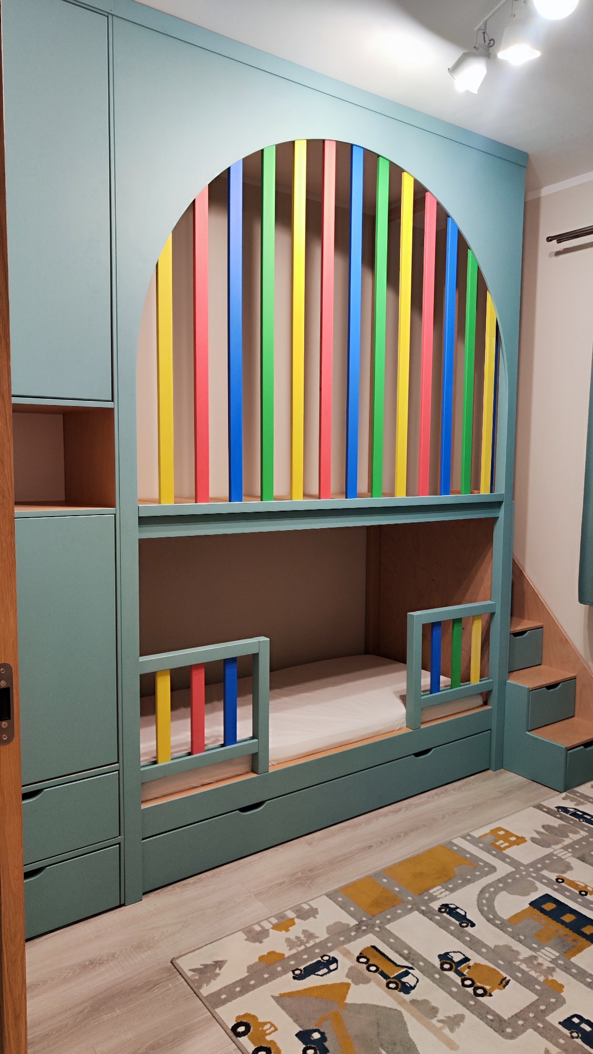 Nietypowe łóżko piętrowe z szafą w kolorze morskim, z kolorowymi szczebelkami i schodkami z szufladami, na tle wzorzystego dywanu w pokoju dziecięcym.