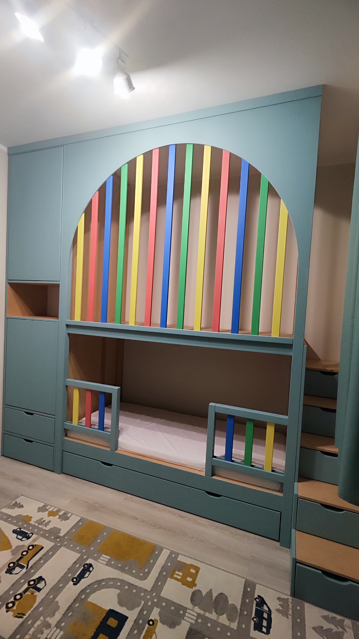 Niebieskie łóżko piętrowe z szafkami i schodami, z kolorowymi szczebelkami w kształcie łuku. Na podłodze dywan z motywem ulic i samochodów.