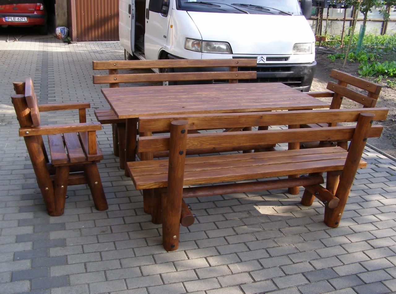 Rustykalny zestaw mebli ogrodowych z ciemnego drewna, składający się ze stołu i ławek z oparciami, ustawiony na brukowanej nawierzchni przed białym samochodem dostawczym.