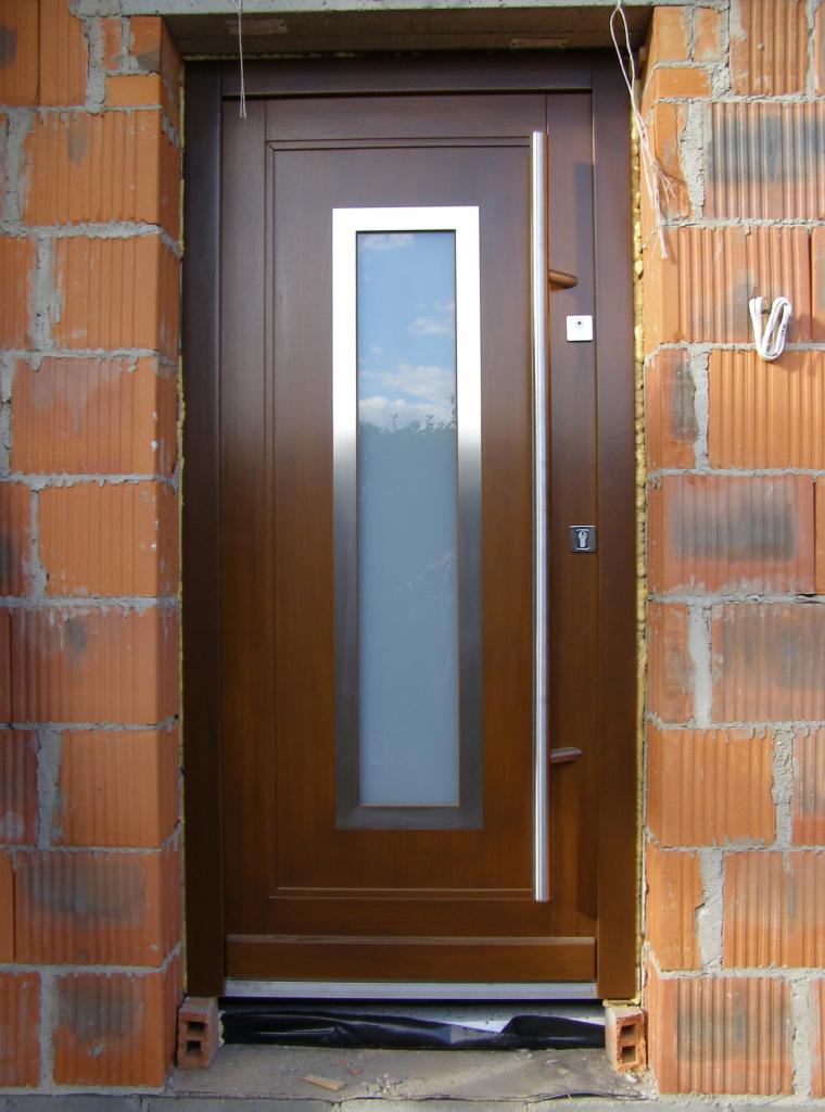 Brązowe drzwi wejściowe z długim, pionowym przeszkleniem w metalowej ramie, zamontowane w surowym murze z czerwonej cegły dziurawki.