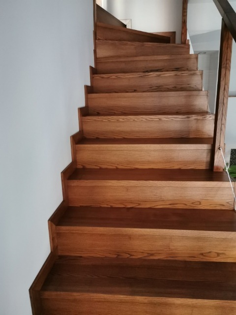 Drewniane schody z ciemnego dębu, widok z dołu, stopnie z wyraźnym usłojeniem, biała ściana po lewej stronie.