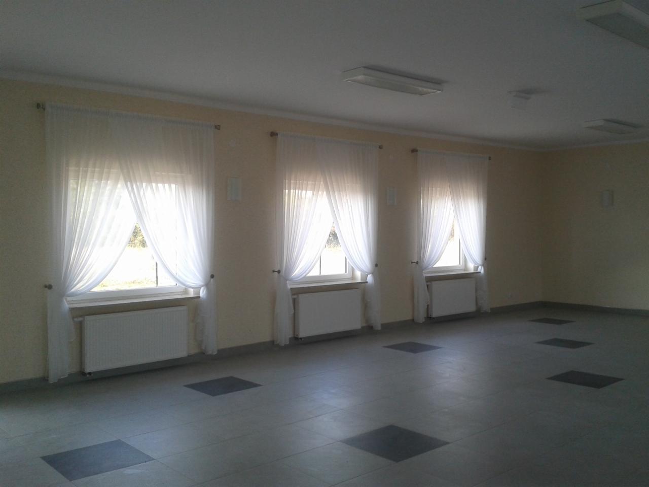 Wnętrze pomieszczenia z trzema oknami zasłoniętymi białymi, zwiewnymi firanami, grzejnikami pod oknami i szarą podłogą wyłożoną kwadratowymi płytkami. Widoczny stan wykończeniowy.