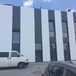 Elewacja budynku biurowego w biało-szare pasy z oknami, częściowo zasłonięta przez samochody, z pracownikiem na dachu.