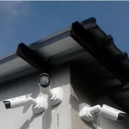 Dwie kamery monitoringu zainstalowane pod okapem budynku, widok z dołu na tle błękitnego nieba.