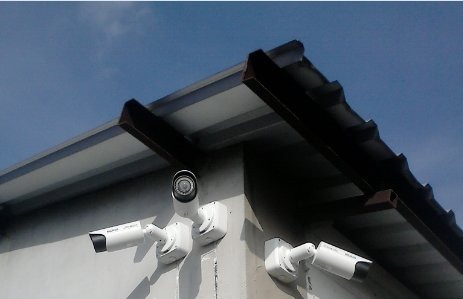 Dwie kamery monitoringu zainstalowane pod okapem budynku, widok z dołu na tle błękitnego nieba.