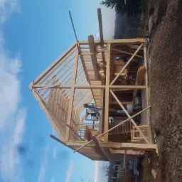 Wznoszenie drewnianej konstrukcji domu w technologii szkieletowej, widoczne elementy więźby dachowej, maszyny budowlane wewnątrz, błękitne niebo w tle.