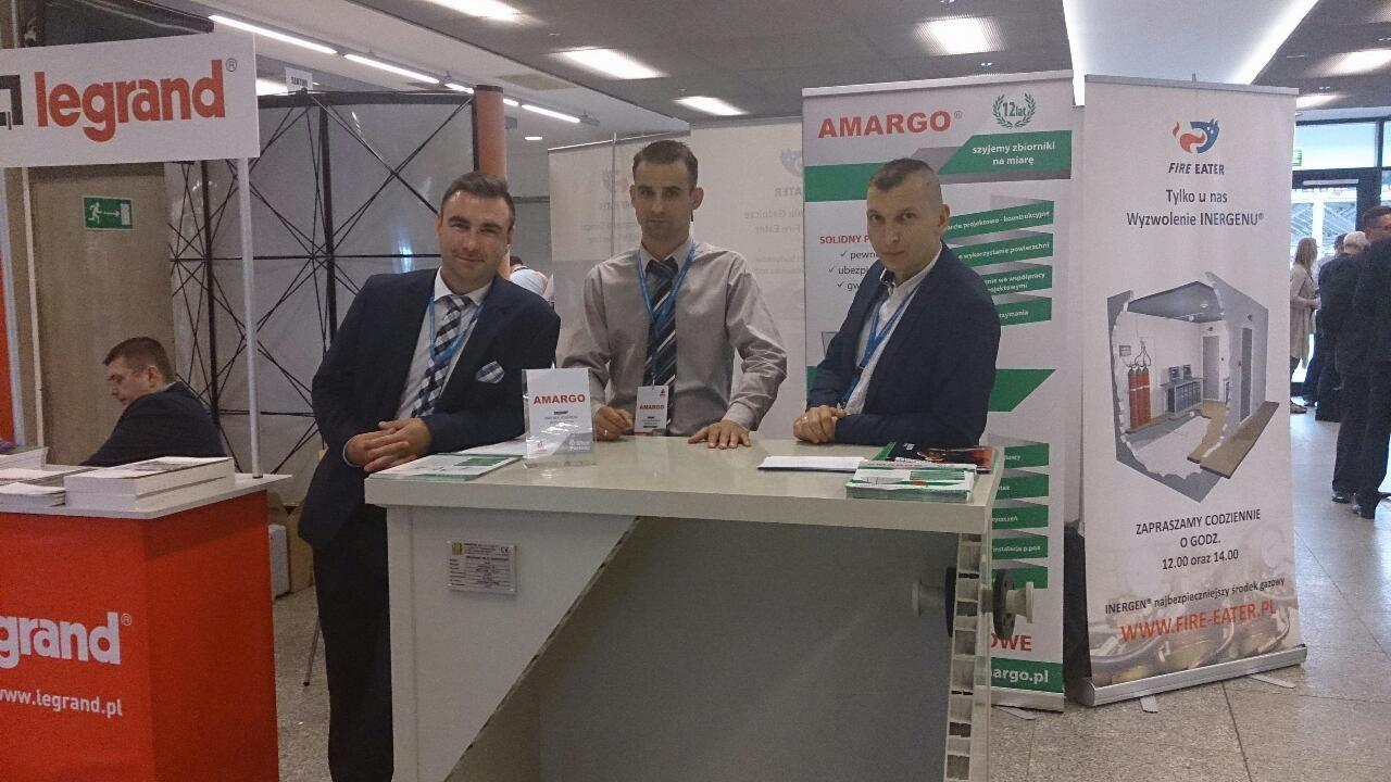 Trzech mężczyzn prezentuje stoisko targowe z logo firm Amargo, Legrand i Fire Eater, oferujących zbiorniki, instalacje i systemy przeciwpożarowe.