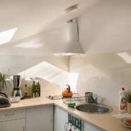 Jasna kuchnia na poddaszu z oknem dachowym, kremowe płytki na ścianie, biały okap, zlewozmywak z suszarką, widoczne naczynia i środki czystości, na blacie przygotowane kanapki z pomidorami.