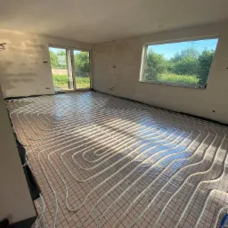 Widok na podłogę z zamontowanym ogrzewaniem podłogowym w jasnym, surowym wnętrzu. Białe rury ułożone w pętle na czerwonej siatce, odbijające światło słoneczne. Okna z widokiem na zieleń na zewnątrz.